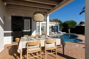 An outdoor dining area with a table set for two, a hanging light fixture, and a swimming pool in the background. - Vrbo Property (Sant Lluís)