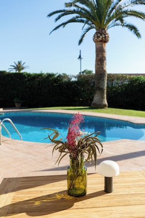 A poolside table with a vase of flowers and a lamp, a palm tree, and a clear blue pool. - Vrbo Property (Sant Lluís)