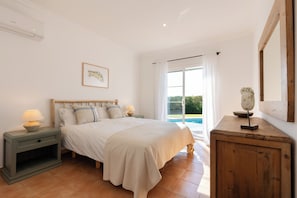 A bedroom with a bed, nightstands, a lamp, a wooden cabinet, a framed picture, and a sliding glass door leading to a pool. - Vrbo Property (Sant Lluís)