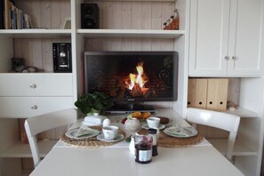 Ein Essbereich mit einem Fernseher, der einen Kamin zeigt, einem weißen Tisch mit Untersetzern, einer Pflanze und einem Marmeladenglas.