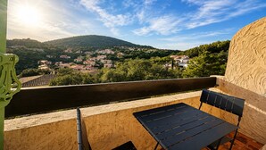 A balcony with a table and chairs, overlooking a village with houses and a hill. - Cosy studio, magnificent swimming pool with exceptional view of the Gulf of St-Tropez (Roquebrune-sur-Argens)