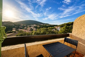 A balcony with a table and chairs, overlooking a village with houses and a hill. - Cosy studio, magnificent swimming pool with exceptional view of the Gulf of St-Tropez (Roquebrune-sur-Argens)