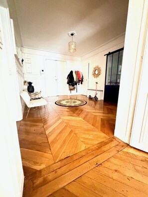 Ein geräumiges Zimmer mit Fischgrätparkett, einem weißen Stuhl, einem Kleiderständer mit mehreren Mänteln und einem Spiegel an der Wand.