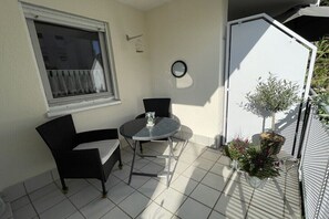 Eine kleine Terrasse im Freien mit einem runden Tisch und zwei Stühlen, einem Fenster mit Vorhängen, einem Blumentopf und einer weißen Tür.