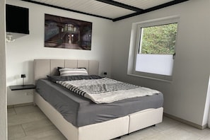 A bedroom with a bed, a nightstand, a wall-mounted TV, a window with blinds, and a painting on the wall. - Holiday apartment with 3 bedrooms (Sinzig)