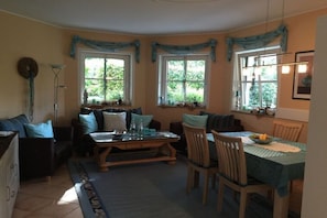 Ein gemütliches Wohnzimmer mit einer Sofagarnitur, einem Esstisch mit Stühlen und einem Fenster mit Vorhängen.