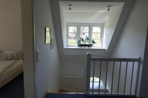 Ein Zimmer mit schrägverdeckter Decke, einem Fenster mit einem Schiffmodell, einem Bett und einem weißen Geländer.