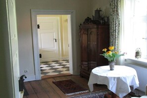 Ein Zimmer mit Schachbrettmuster-Boden, einer weißen Tür, einem Holzschrank, einem Tisch mit weißem Tuch und einer Vase mit gelben Blumen.