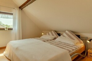 Une chambre avec un plafond en pente, un lit avec une literie à rayures, une lampe de chevet et une fenêtre avec un rideau.