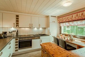 Une cuisine avec des meubles blancs, une table en bois et une fenêtre agrémentée d’un lambrequin à carreaux.