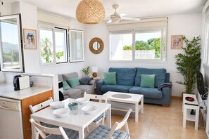 Ein modernes Wohnzimmer mit einer Couch, einem Esstisch und Stühlen, einer Küchenzeile mit Kaffeemaschine und großen Fenstern mit Blick auf Grünflächen.