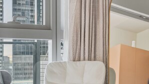 Une pièce moderne avec un fauteuil blanc, une table ronde en bois, un grand miroir et une vue sur les bâtiments de la ville, visible par la fenêtre.