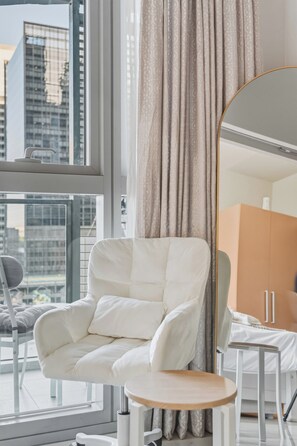 A modern room with a white armchair, a round wooden table, a large mirror, and a view of city buildings through a window. - Luxury BGC Condo • Hotel Vibes • Netflix+Workspace (Taguig)