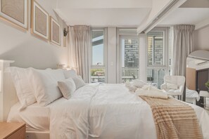 A hotel room with a large bed, a chair, a desk, and a view of the city through large windows. - Luxury BGC Condo • Hotel Vibes • Netflix+Workspace (Taguig)