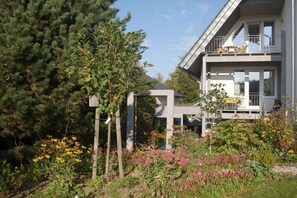 Een modern herenhuis met twee verdiepingen en een balkon, omgeven door een tuin vol planten en bloemen.