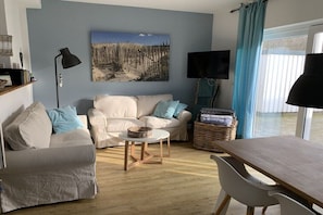Ein Wohnzimmer mit einer Couch, einem kleinen runden Tisch, einem Fernseher und einem Fenster mit Vorhängen.