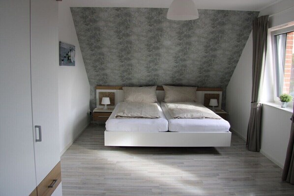 A modern bedroom with a sloped ceiling, a large bed, bedside tables, and a window with curtains. - Holiday home with 4 bedrooms (Niedersachsen - Garrel)