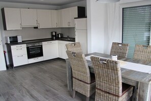 A modern kitchen with white cabinets, a built-in oven, and a dining area with wicker chairs and a wooden table. - Holiday home with 4 bedrooms (Niedersachsen - Garrel)