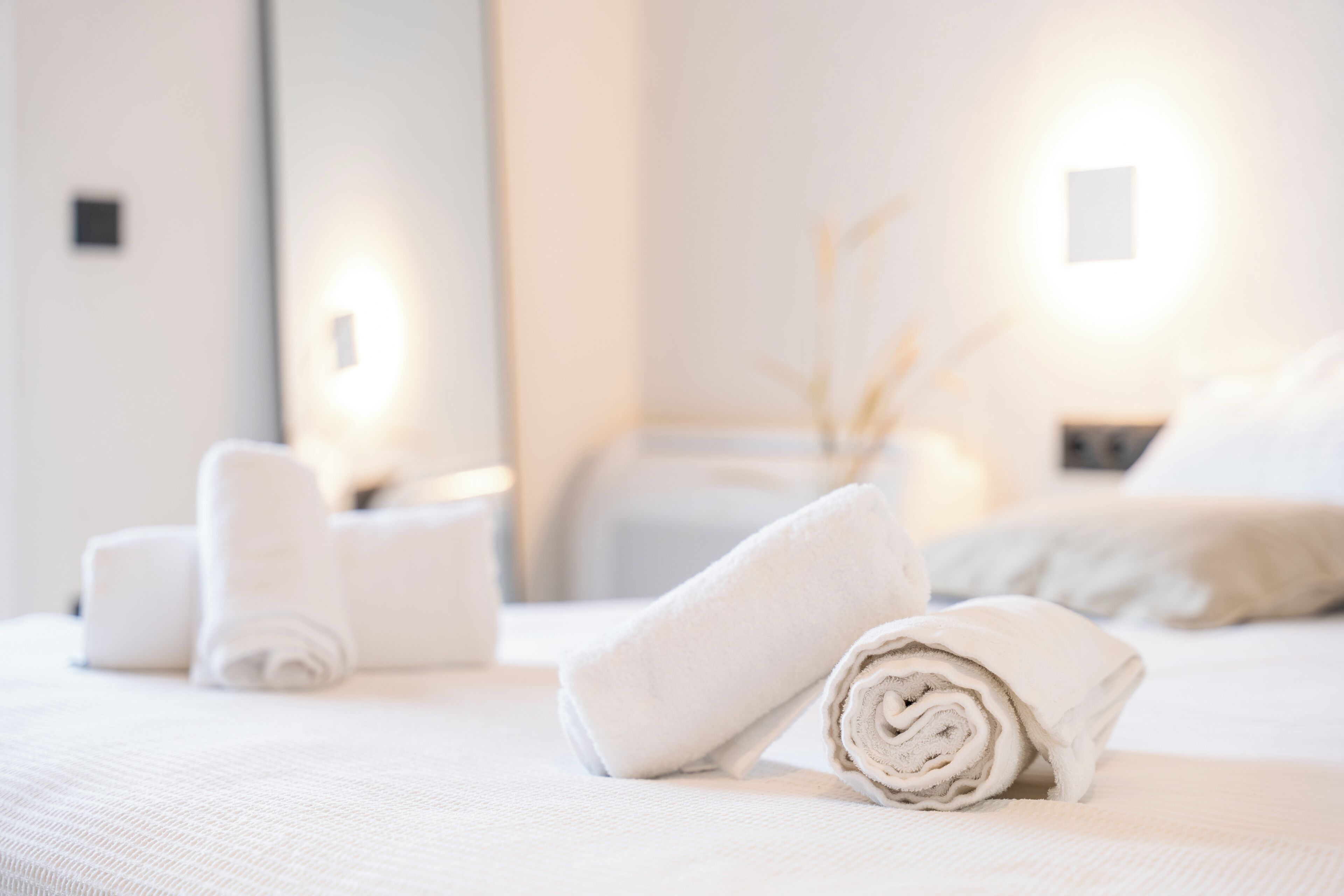 Ein ordentlich bezogenes Bett mit aufgerollten weißen Handtüchern, ein Nachttisch mit Lampe und eine Wandleuchte.