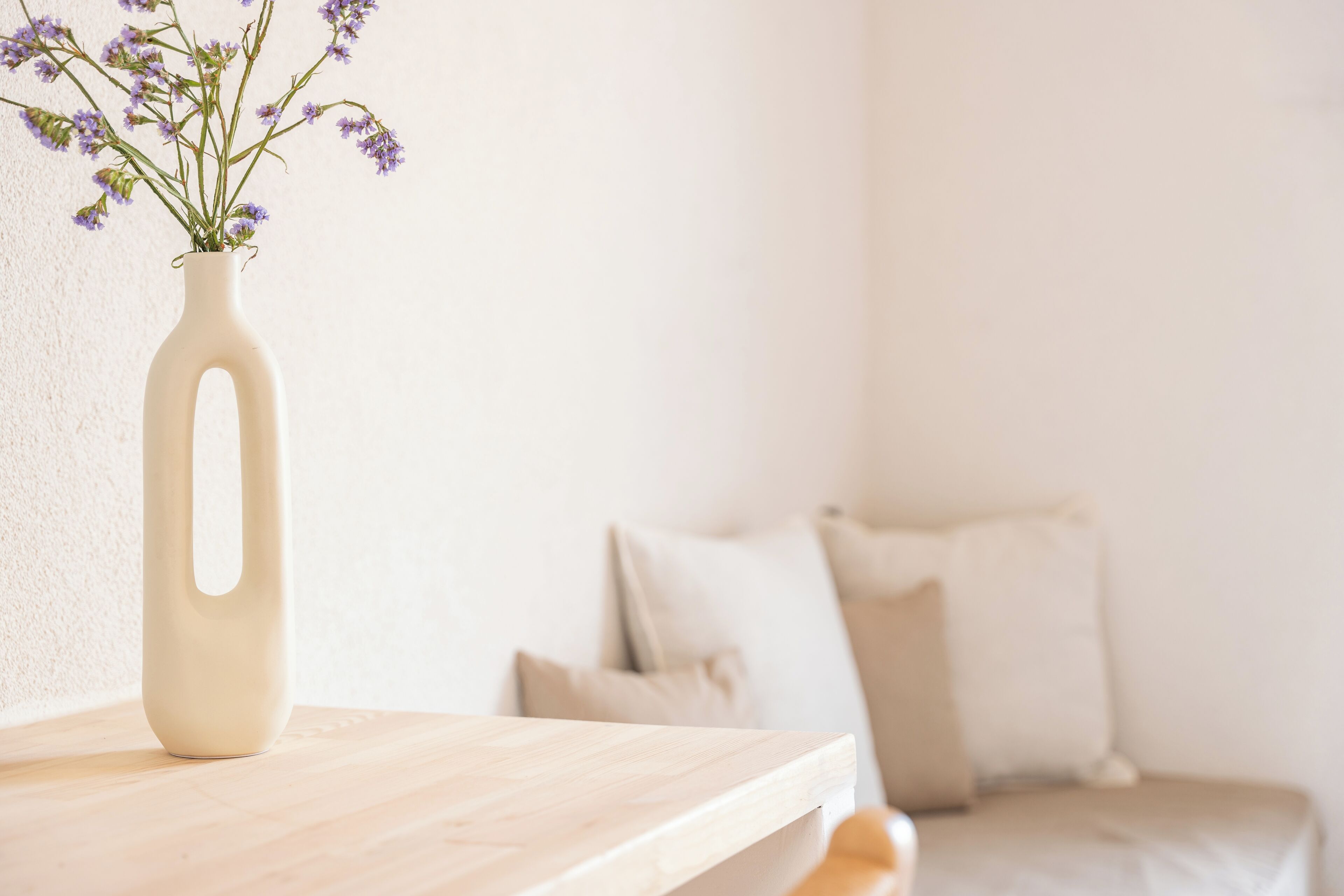 A beige vase with purple flowers on a wooden surface, a bed with beige bedding, and a plain wall in the background.