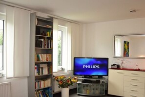 Ein Wohnzimmer mit Fernseher, Bücherregal, weißem Schrank und Fenster.