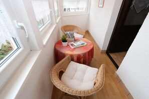 Ein schmales Zimmer mit einem Korbsessel, einem kleinen Tisch mit roter Tischdecke und einem Fenster mit Blick.