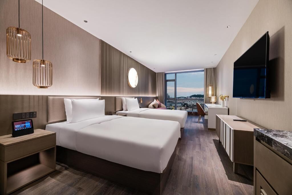 A modern hotel room with two beds, a TV, a desk, and a view of the city.