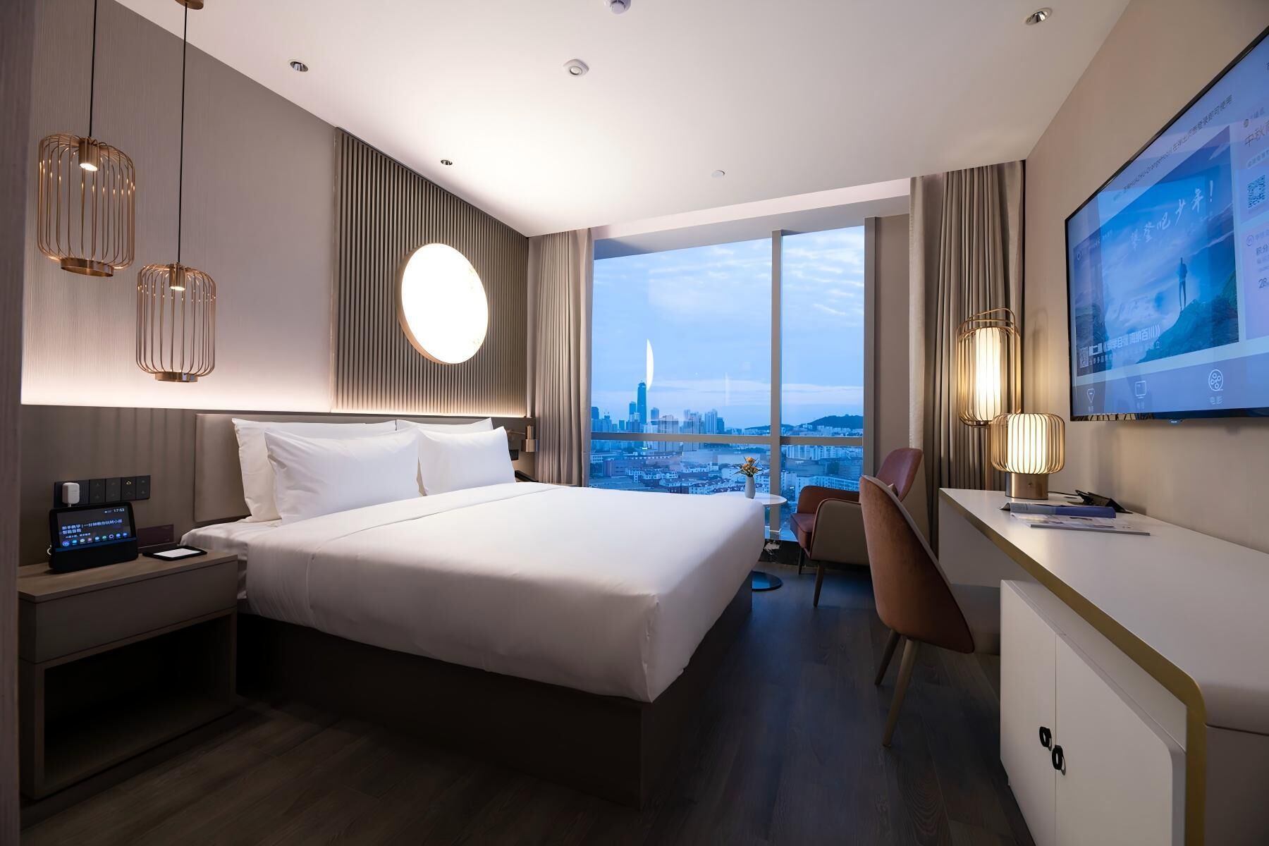 A hotel room with a bed, bedside table, desk, chair, TV, and a city view.