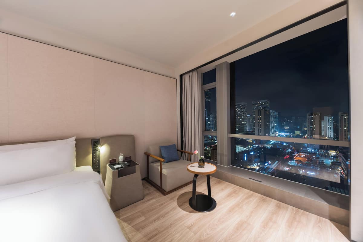 Quarto de hotel moderno com uma janela grande, uma cama, uma mesa pequena e uma mesinha de cabeceira.