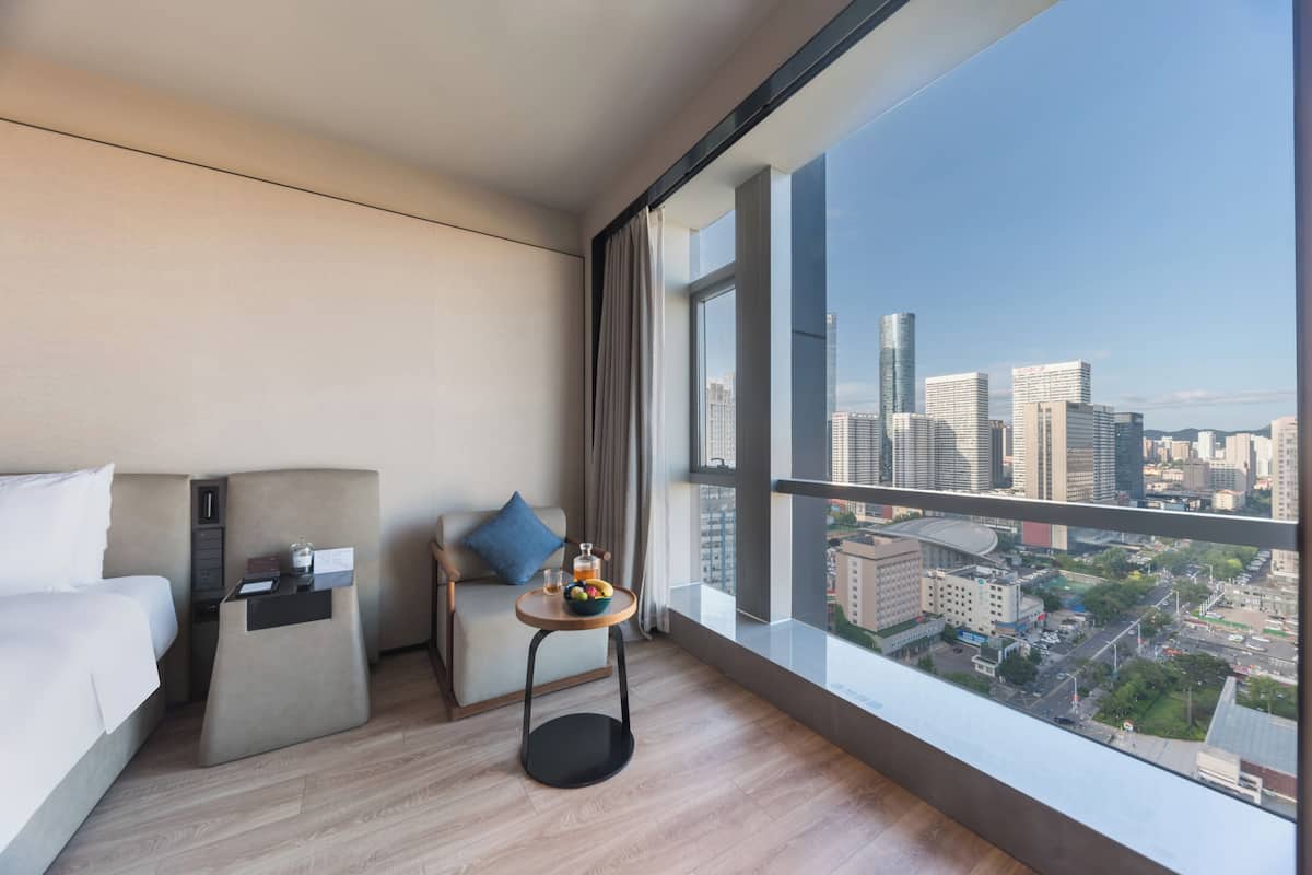 Quarto de hotel moderno com uma janela grande, uma cama, uma mesinha com frutas e vista para a paisagem urbana.