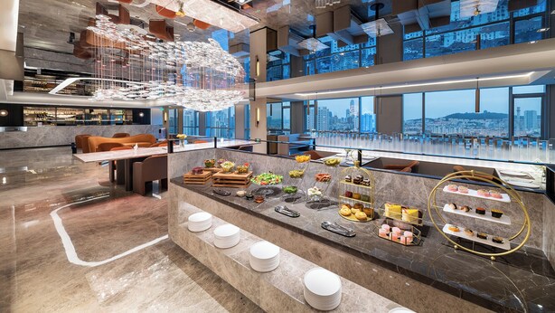 A modern hotel lobby with a marble reception desk, a variety of food and drink options, and a city view through large windows. - Crystal Hotel Qingdao MixC Shandong Road (Qingdao)