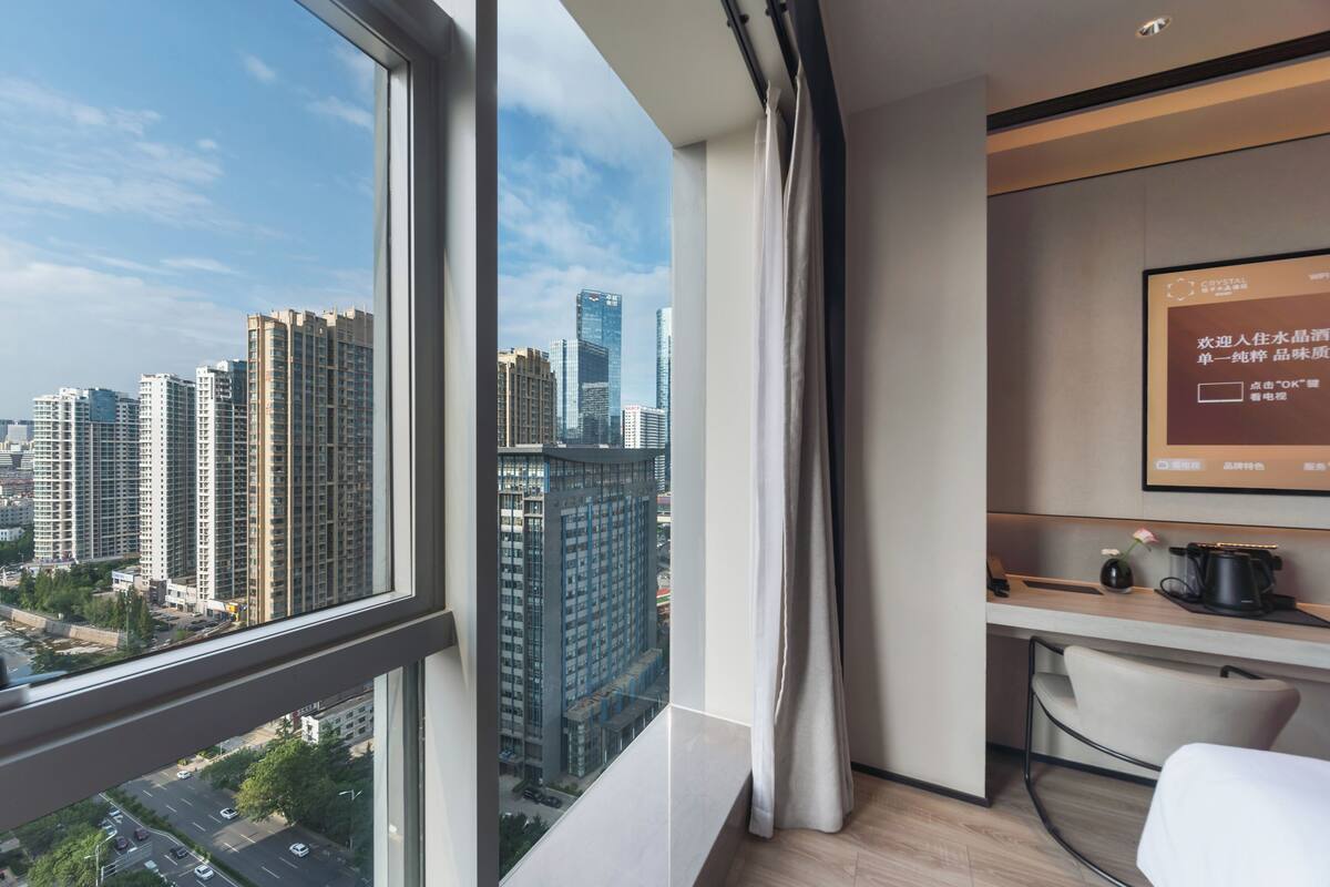 Quarto de hotel moderno com uma janela grande que oferece vista da cidade, uma escrivaninha com cadeira e uma tela montada exibindo informações.