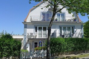 Ein zweistöckiges Haus mit Balkon, weißem Zaun und einem Baum im Vordergrund.