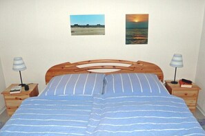 Ein Schlafzimmer mit einem hölzernen Bett, gestreifter Bettwäsche, zwei Nachttischlampen, einem Nachttisch mit Buch und Fernbedienung sowie zwei gerahmten Bildern an der Wand.