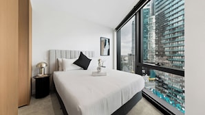 Un hotel moderno con una cama grande, una mesita de noche, una lámpara y vistas a un paisaje urbano a través de un ventanal amplio.
