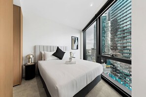 Ein modernes Hotelzimmer mit einem großen Bett, einem Nachttisch, einer Lampe und Blick auf eine Stadtlandschaft durch ein großes Fenster.