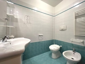 Ein Badezimmer mit einem weißen Waschbecken, einer Toilette und einem Bidet. Die Wände sind in einem zweifarbigen Muster gefliest, und es gibt eine Duschkabine aus Glas.