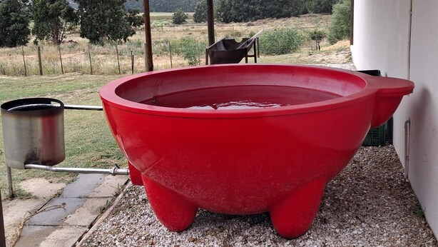 A red hot tub with a metal container beside it, set on a gravel surface with a view of a rural landscape.