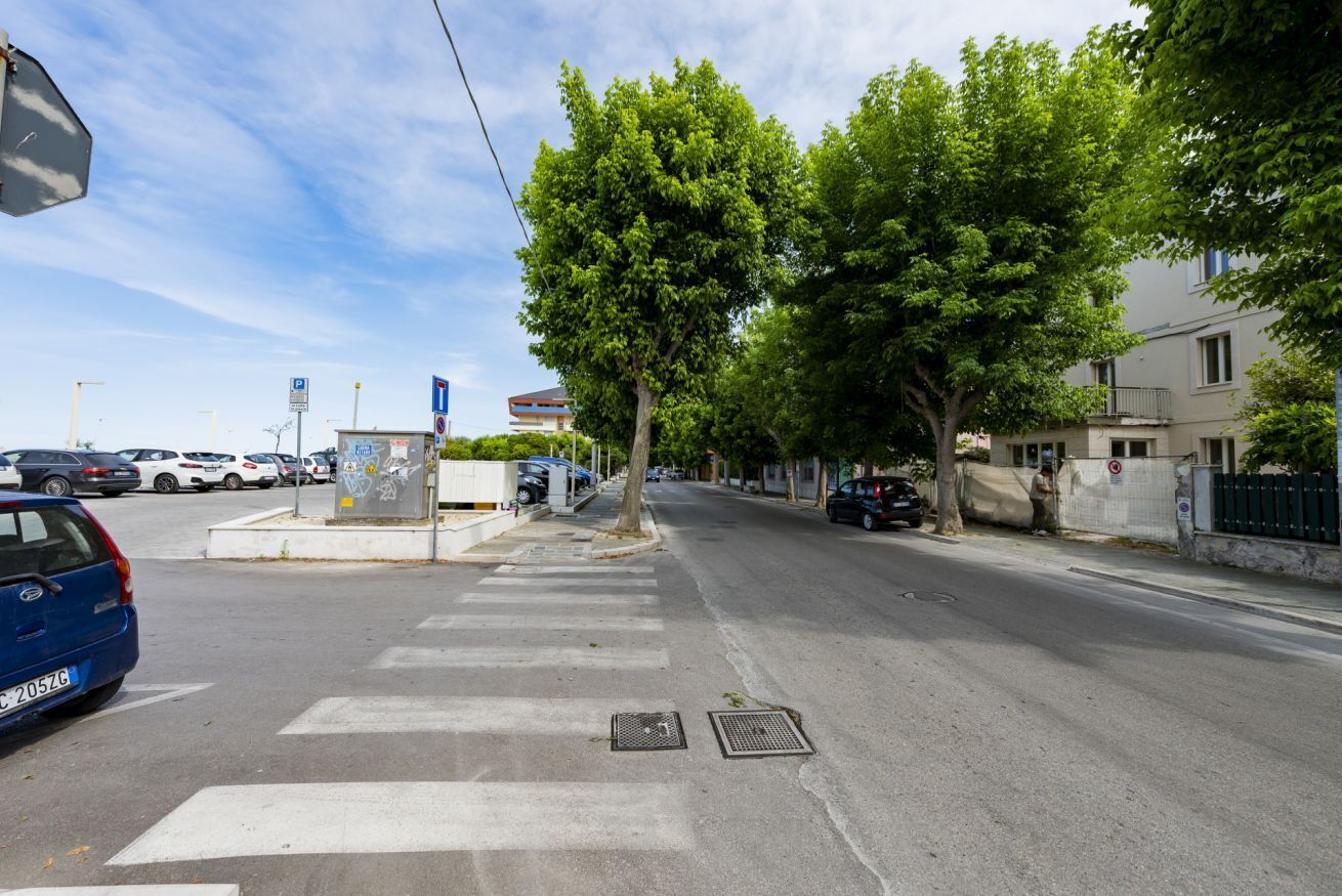 A street with a pedestrian crossing, trees, parked cars, and a building with a balcony.