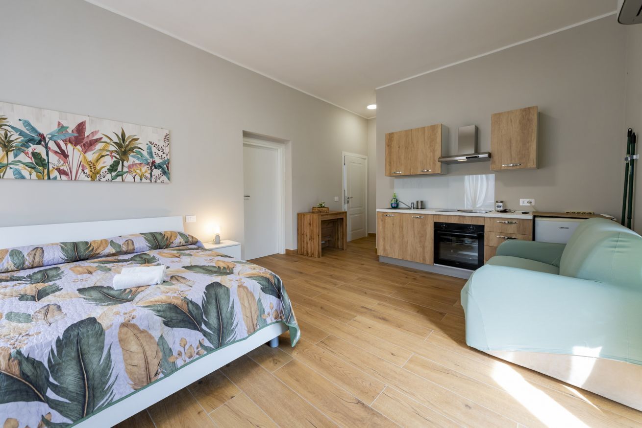 A bedroom with a bed, a nightstand, a painting, a kitchenette with cabinets, a stove, and a sofa.