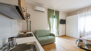 A kitchenette with a sink, a stove, and a microwave. There is a green sofa and a bed with a patterned blanket. An air conditioning unit is mounted on the wall. - GuestHost - Cozy Balcony Flat X3 near the Sea (Francavilla al Mare)