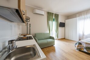 Angolo cottura con lavandino, fornello e microonde. Un divano verde e un letto con coperta fantasia. Un condizionatore d'aria è montato sulla parete.