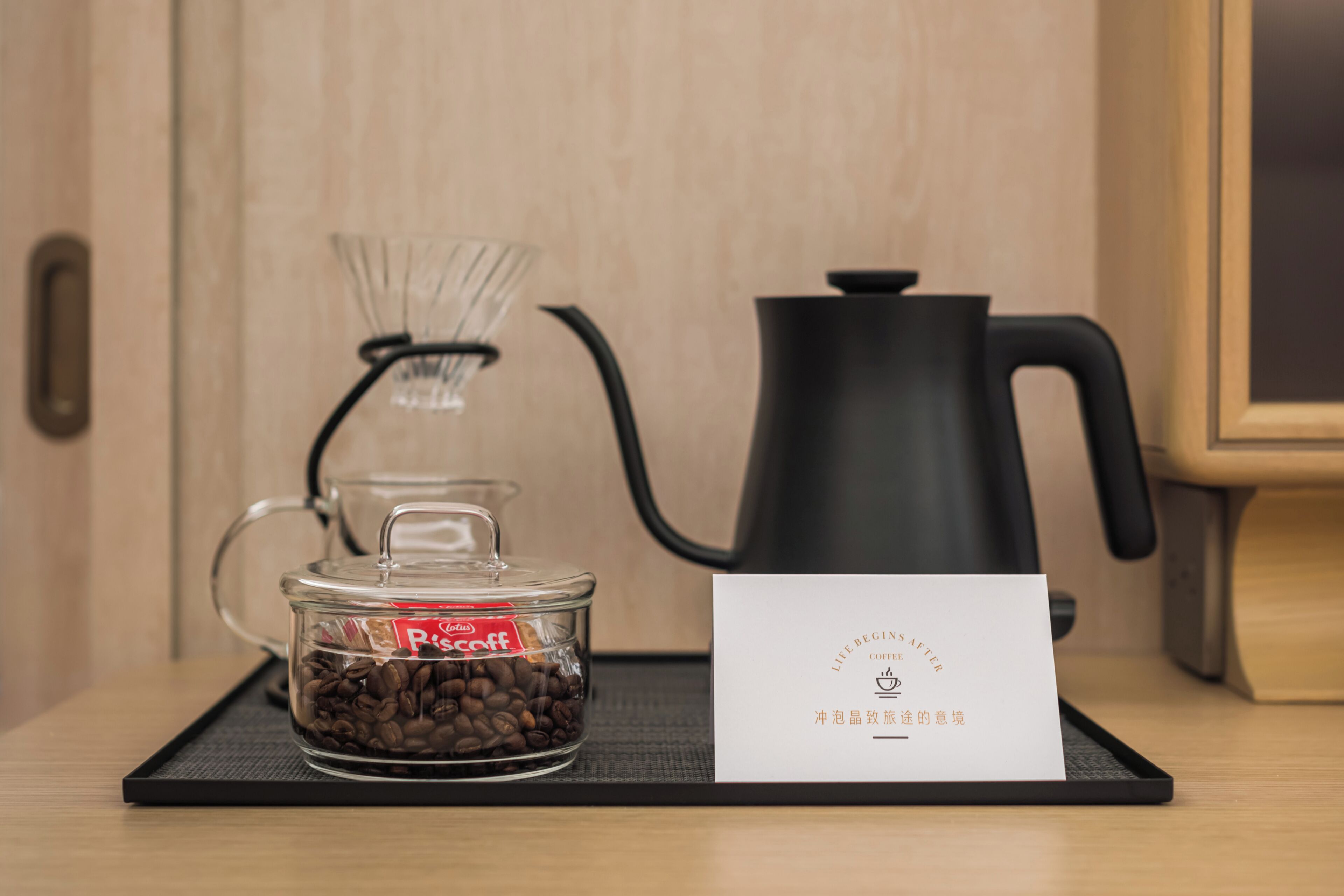 A coffee maker, a glass container with coffee beans, and a card on a tray.