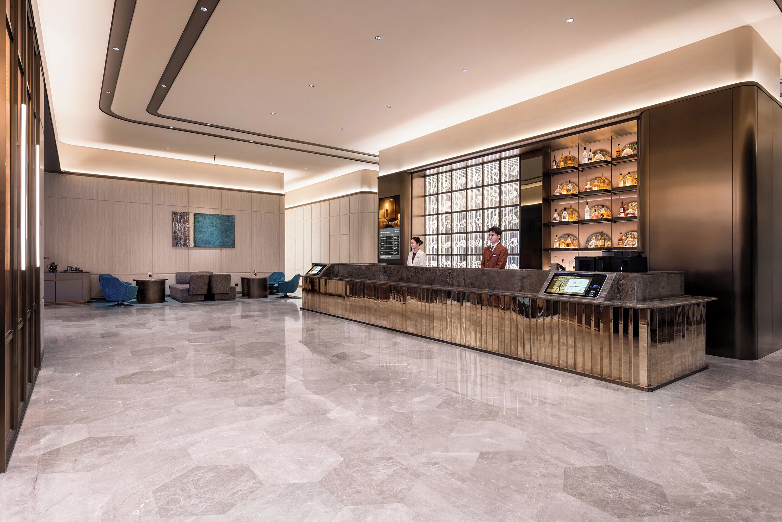 A modern hotel lobby with a reception desk, seating area, and a wall of decorative bottles.