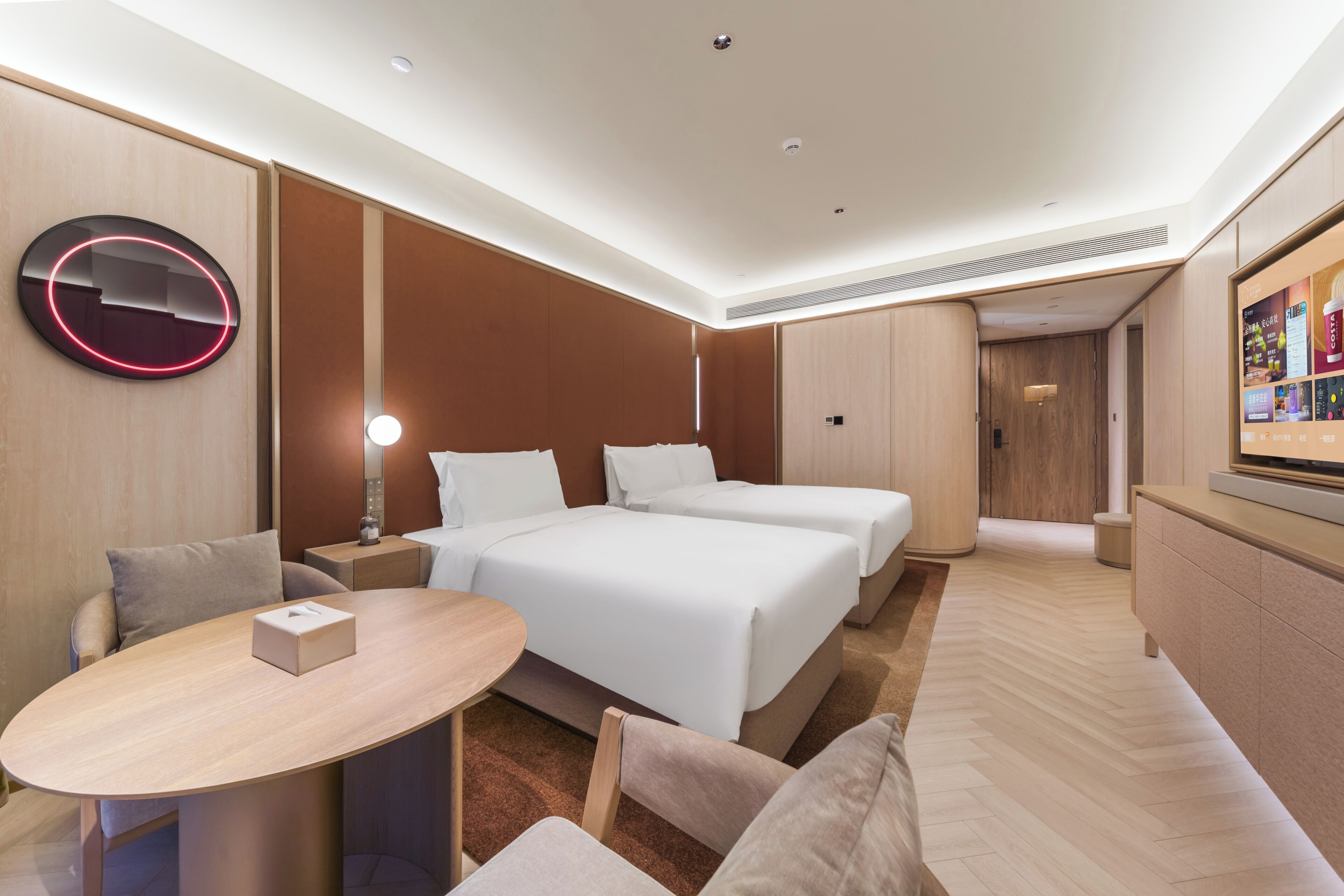 A hotel room with two beds, a round table, a sofa, a TV, and a wall-mounted circular light.