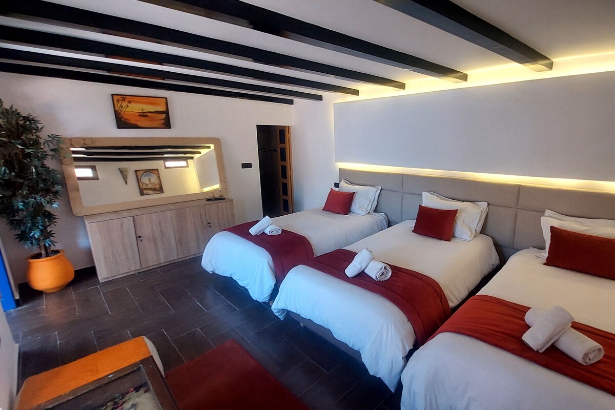 A hotel room with three beds, a small desk, a potted plant, and a painting on the wall.