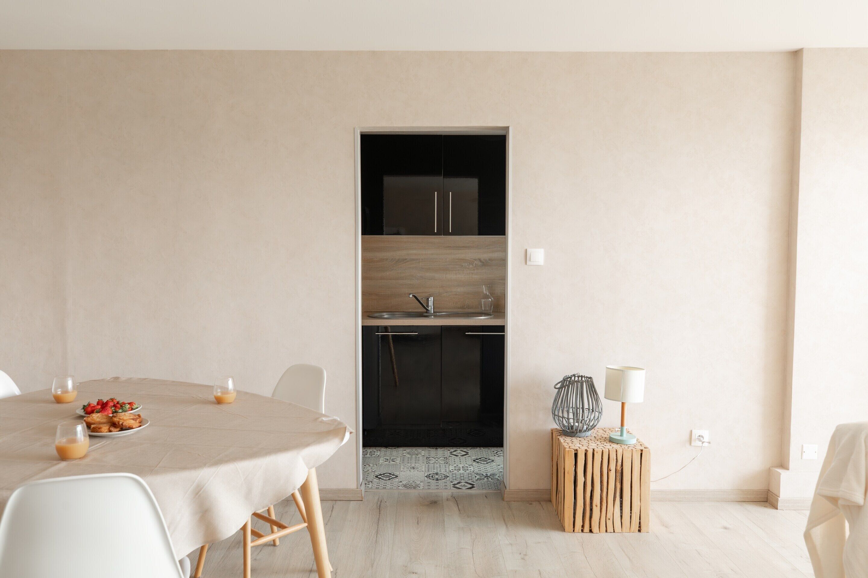 A modern kitchen with a dark countertop and cabinets, a small dining table with chairs, and a decorative lamp on a wooden stool.