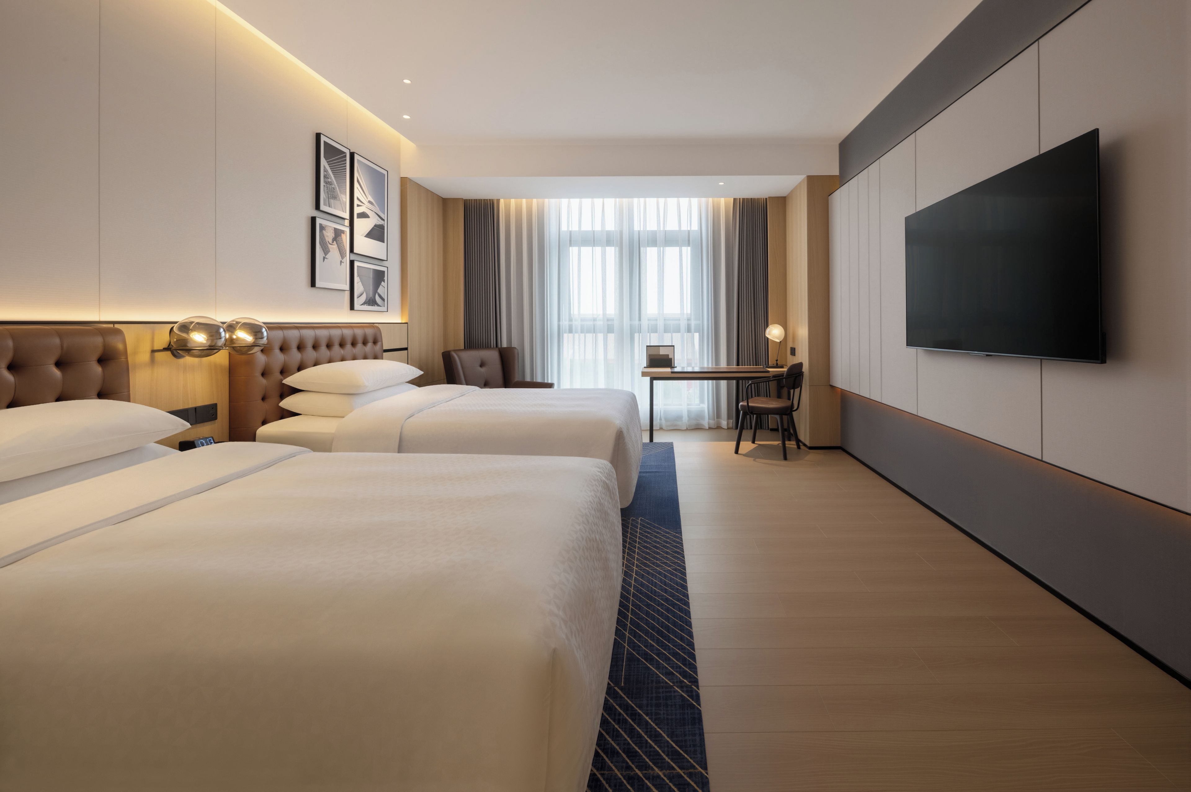 A hotel room with two beds, a desk, a chair, a flat-screen TV, and a large window with curtains.
