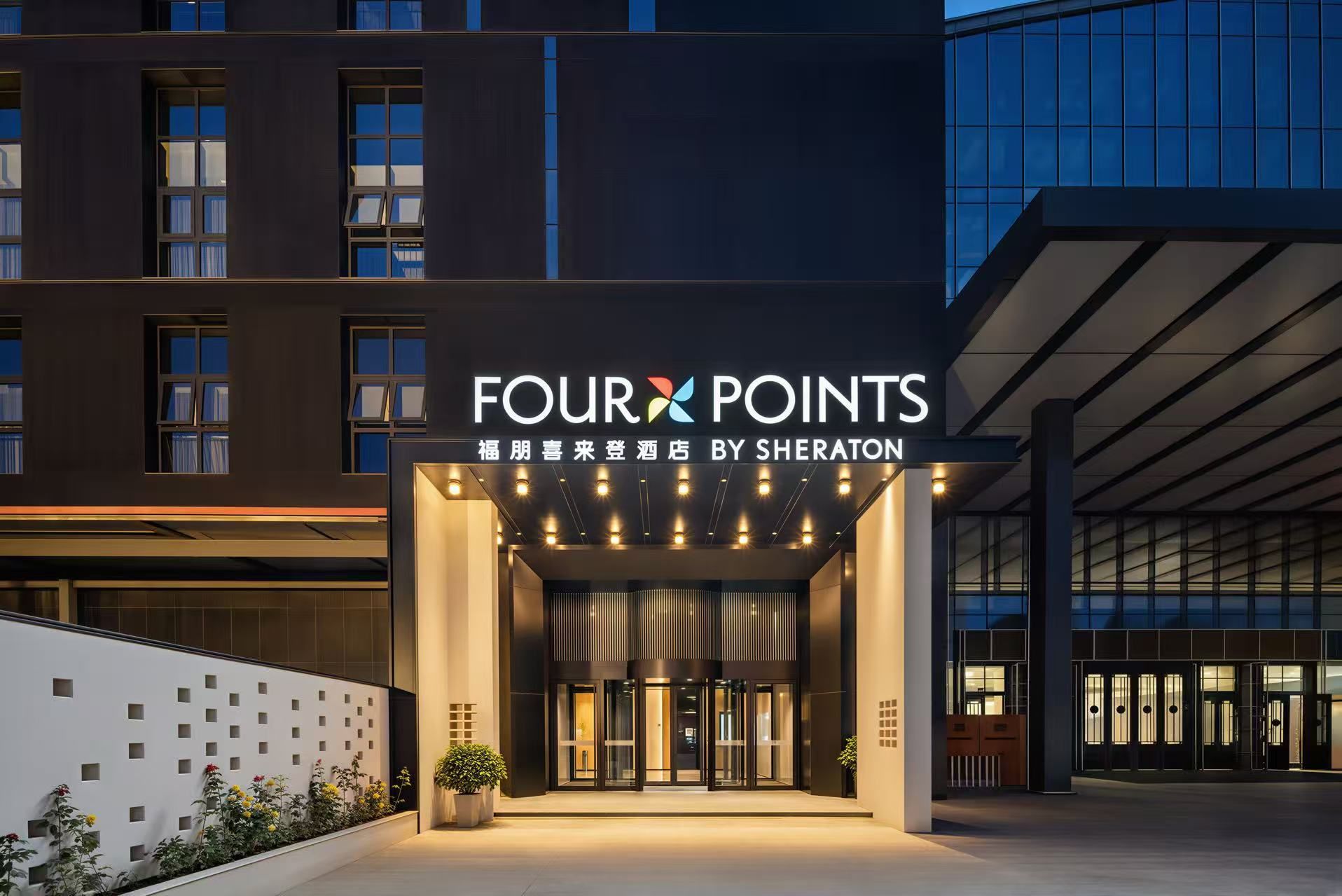 Entrada de un hotel con un cartel que dice 'FOUR POINTS BY SHERATON' y una fachada de edificio moderna al fondo.