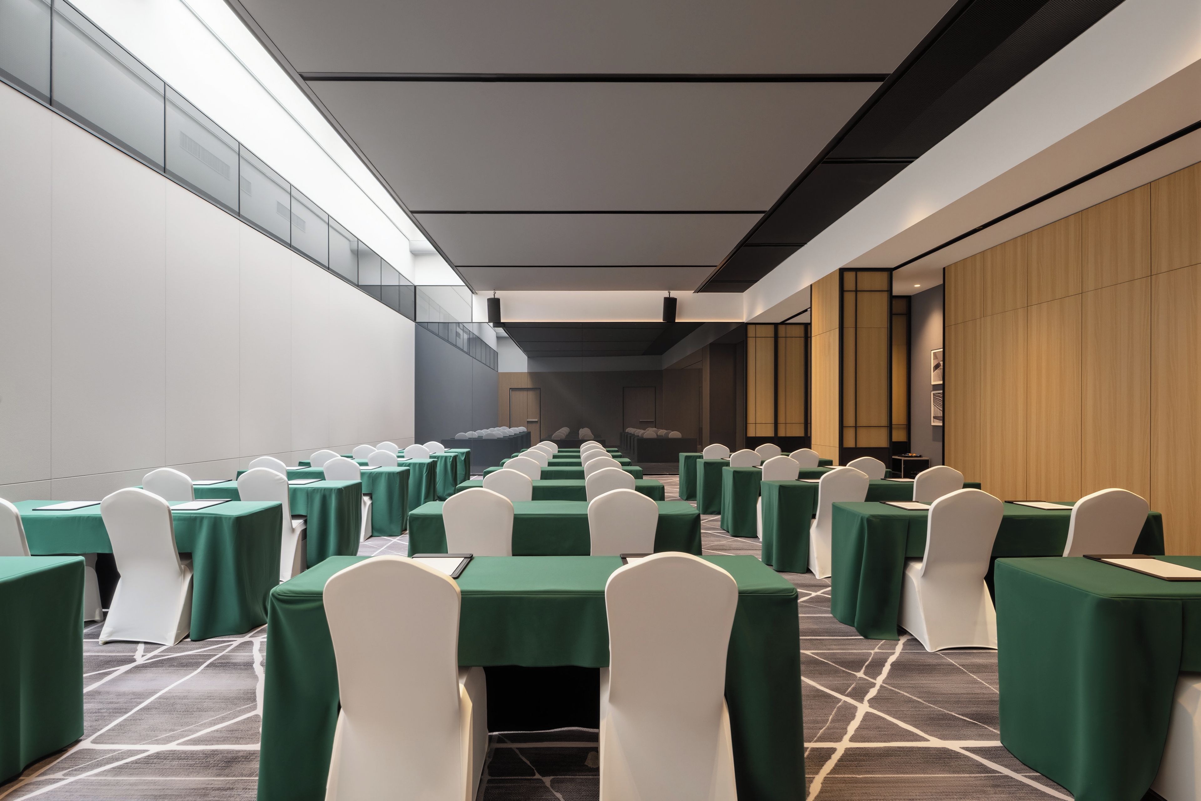 A conference room with rows of tables and chairs, a modern ceiling design, and a view of the adjacent room.